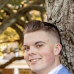 A portrait of Micah Long standing against a tree and looking toward the viewer's left.
