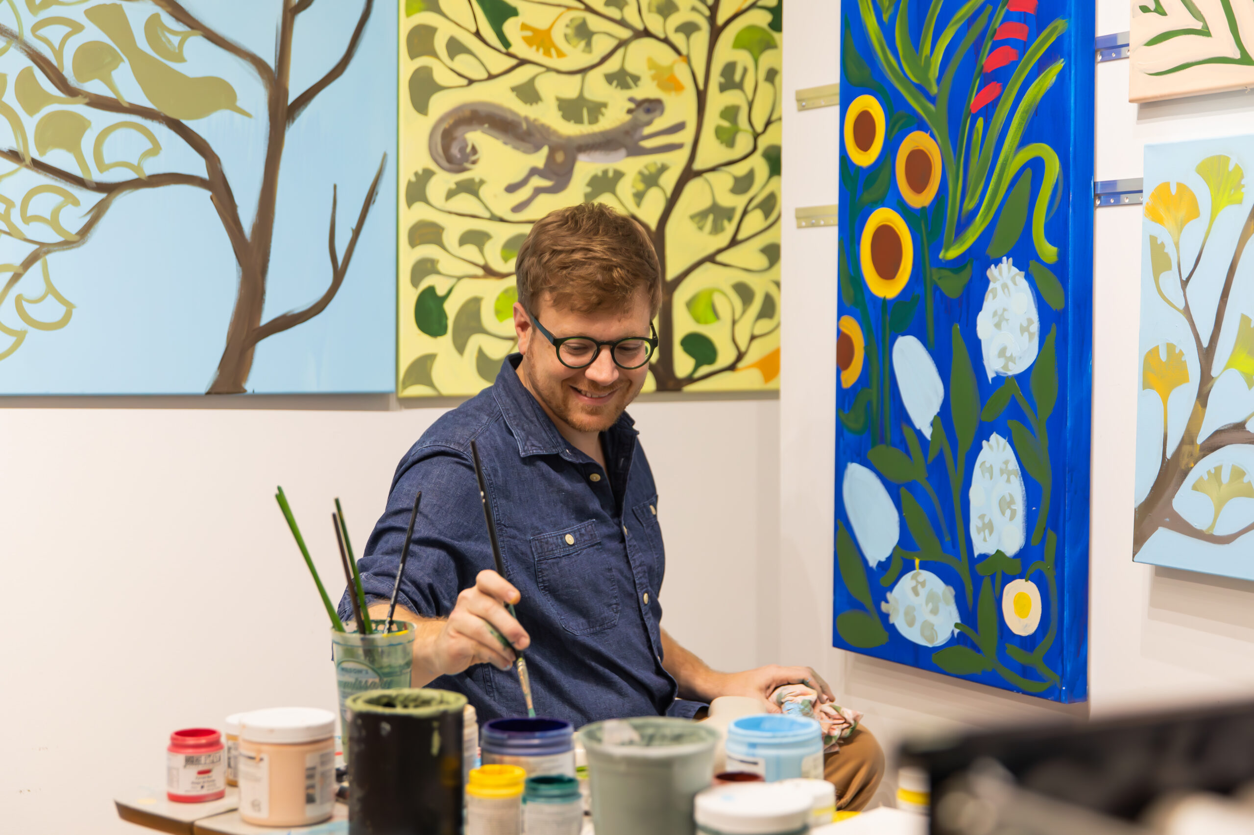 Adam Trest, artist, sitting in his studio with paints on a desk and brightly colored paintings all around while he works on sunflowers on his easel.