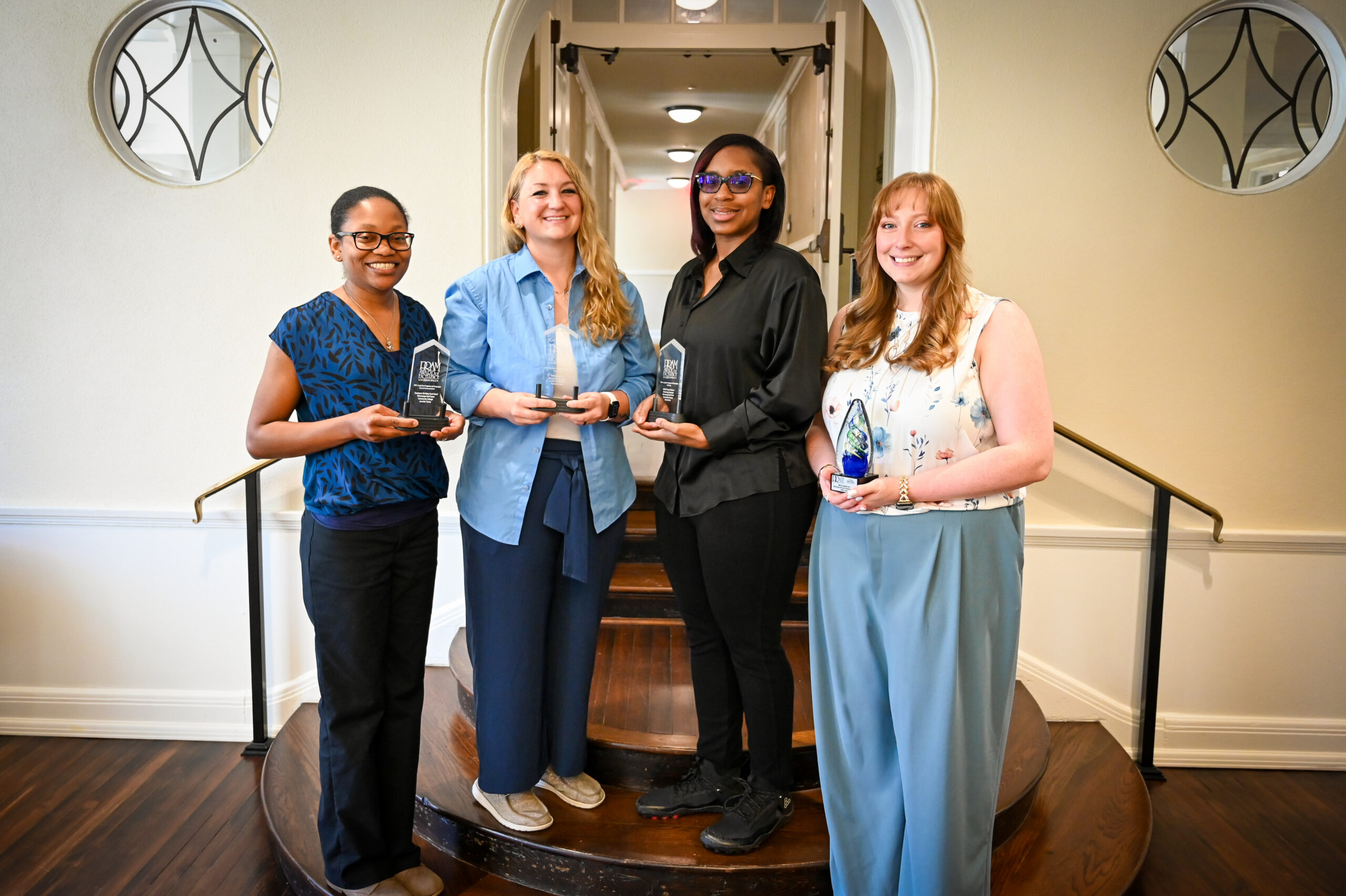 From left standing on stairs with trophies in their hands are Jennifer Tinnin, Creative Officer, Award of Excellence; Elizabeth Brown, Communications & Marketing Specialist, Award of Excellence; Aleá Landry, Graphic Designer, Award of Excellence; and Ashlyn Levins, Chief Creative Officer, PRISM Award.